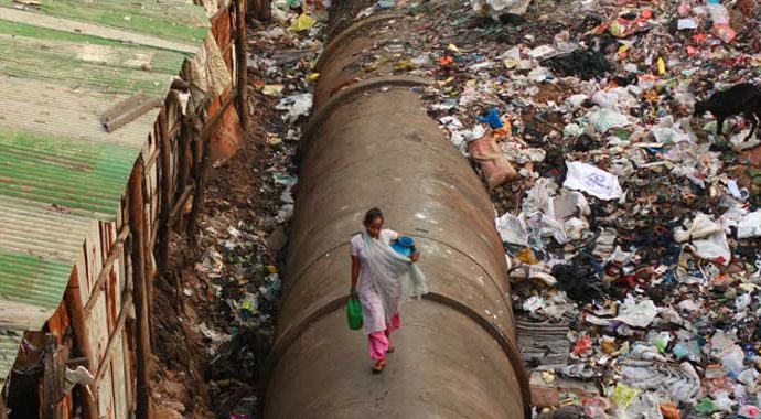 A woman walks along a pipe with a bucket in her hand.