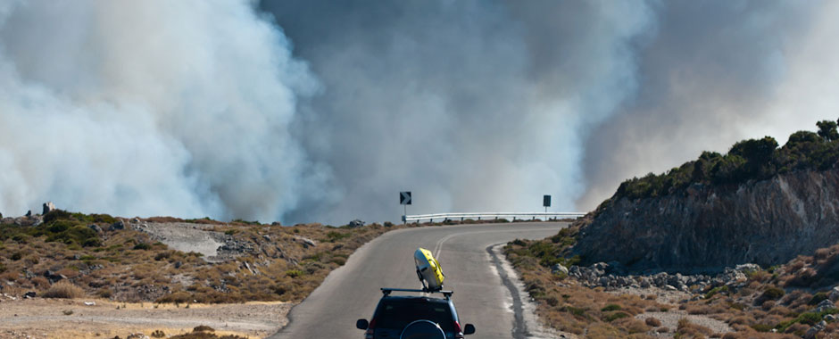 A car drives on a mountain road with clouds of smoke in the background